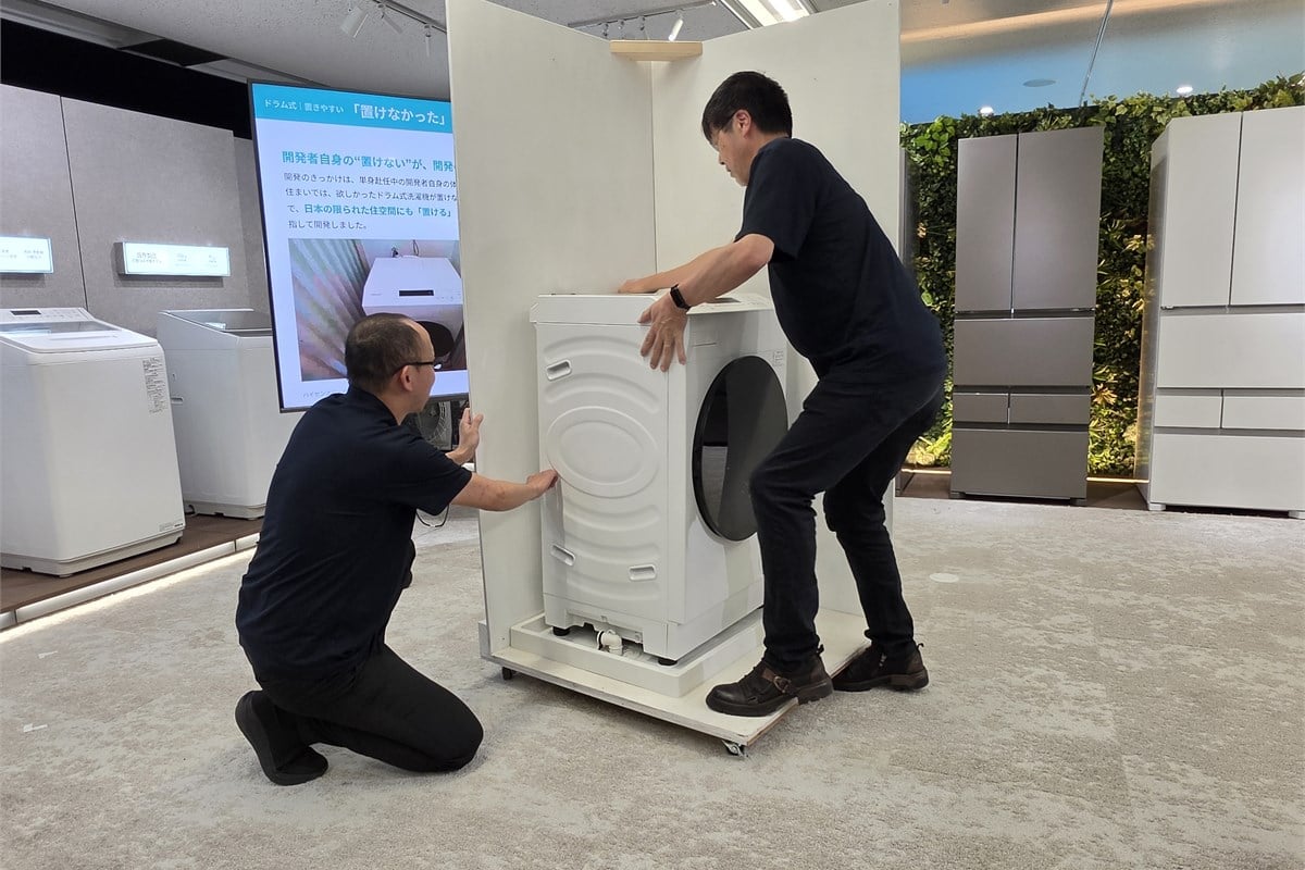 開発者の住まいを参考に設けられた展示スペースへ、新型ドラム式洗濯乾燥機を設置する実演。給水栓や排水口との干渉を避けるための工夫が、数字上の薄型化だけではないことを伝えている。