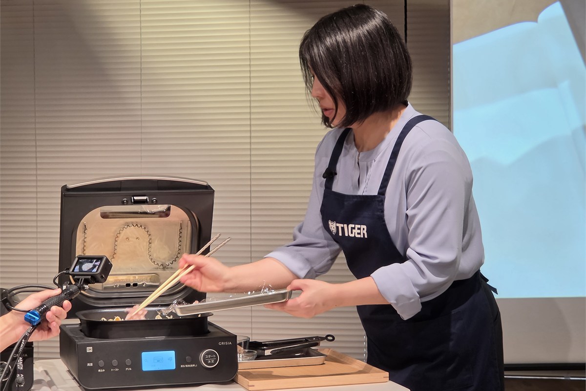 焼きそばの実演で、タイガー魔法瓶の前田真矢子氏が食材をインナーポットに入れる様子。加熱前に材料を重ねるだけで、調理中に混ぜ続ける必要がない。「作る人がコンロに張りつかなくていい」という価値が伝わる場面だ。